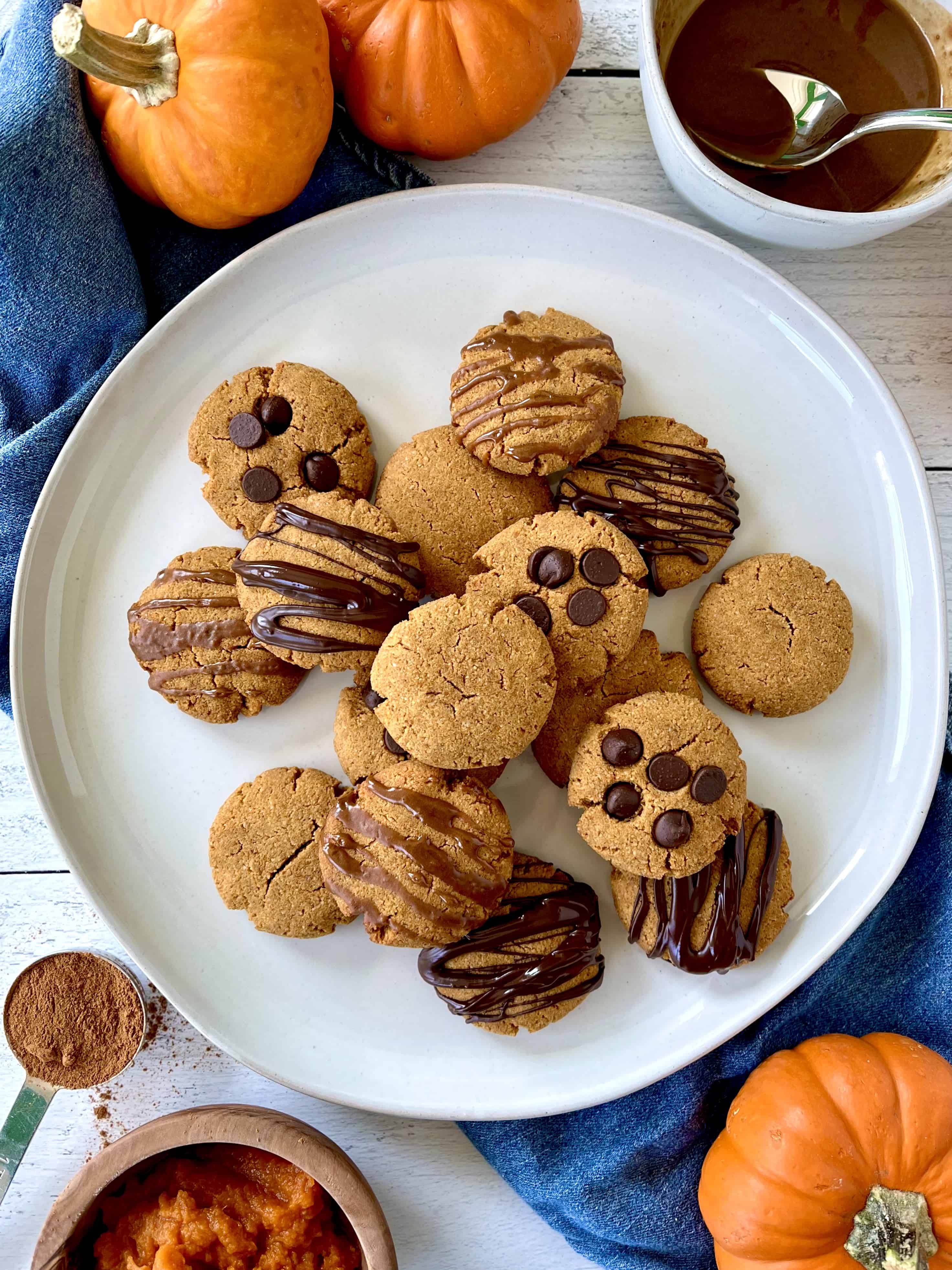 Vegan pumpkin cookies piled on a plate, some with chocolate chips, some with a chocolate or caramel drizzle, some plain.