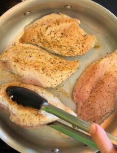A hand using tongs to flip seasoned chicken breasts in a stainless steel pan.