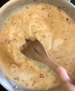 A hand holding a wooden spatula as it stirs a creamy sauce in a stainless steel pan.