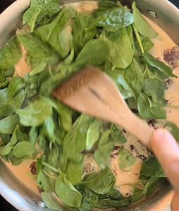 A hand holding a wooden spatula as it stirs fresh spinach leaves into a creamy sauce in a pan.