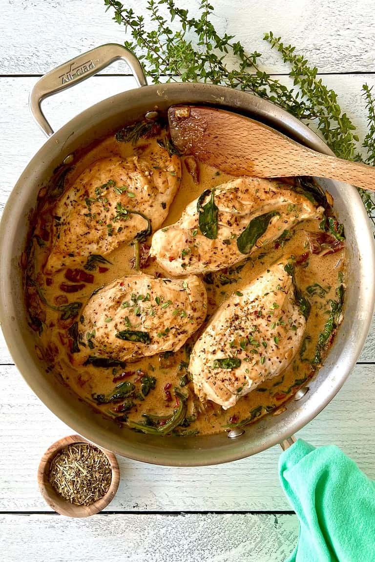 Dairy-free Tuscan chicken in a stainless steel pan with a wooden spatula and a green napkin wrapped around its handle, next to a bunch of thyme sprigs.