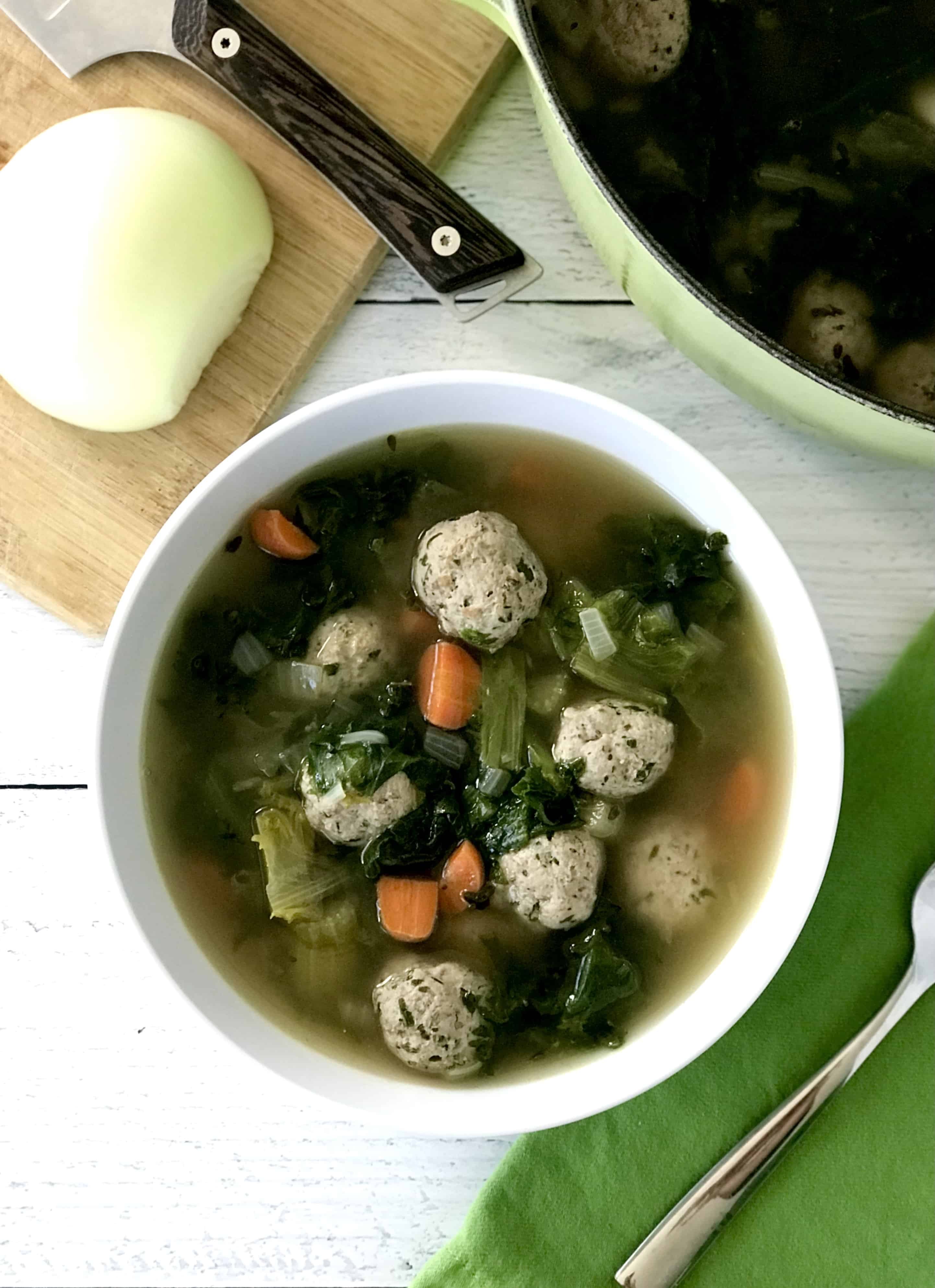 Gluten-Free Italian Wedding Soup in a white bowl next to a green napkin with a spoon on it and an onion on a cutting board.