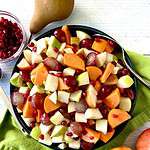 An apple and pear fruit salad in a bowl with a serving spoon, surrounded by an apple, a pear, and orange half and a bowl of pomegranate seeds.