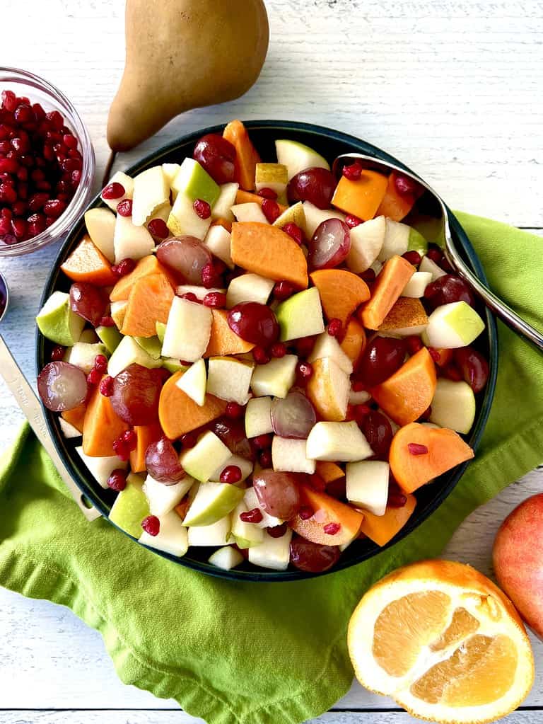 An apple and pear fruit salad in a bowl with a serving spoon, surrounded by an apple, a pear, and orange half and a bowl of pomegranate seeds.