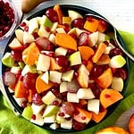 A fruit salad for Thanksgiving in a blue bowl surrounded by an apple, a pear, an orange half and a bowl of pomegranate seeds.