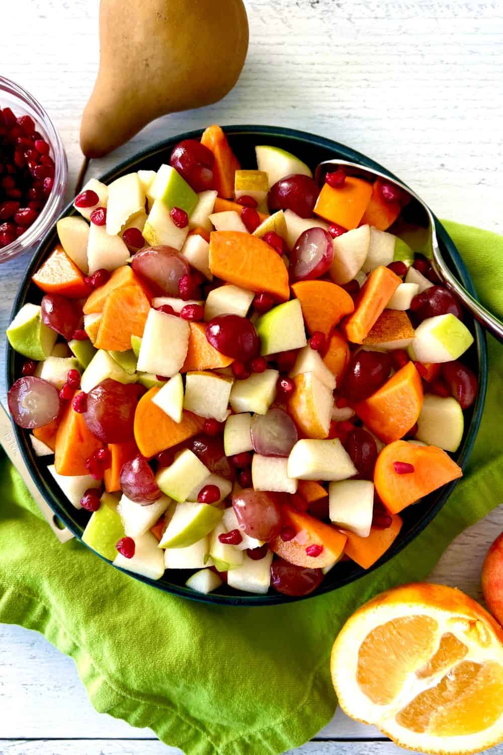 A fruit salad for Thanksgiving in a blue bowl surrounded by an apple, a pear, an orange half and a bowl of pomegranate seeds.
