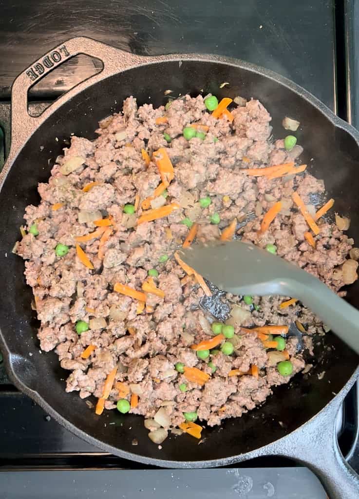 Stirring ground beef, peas and carrots in a cast iron skillet.