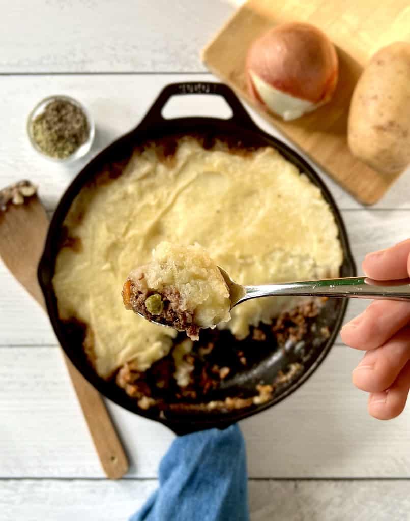 A healthy cottage pie in a cast iron skillet with a hand holding a spoon above it with some of the pie on the spoon.
