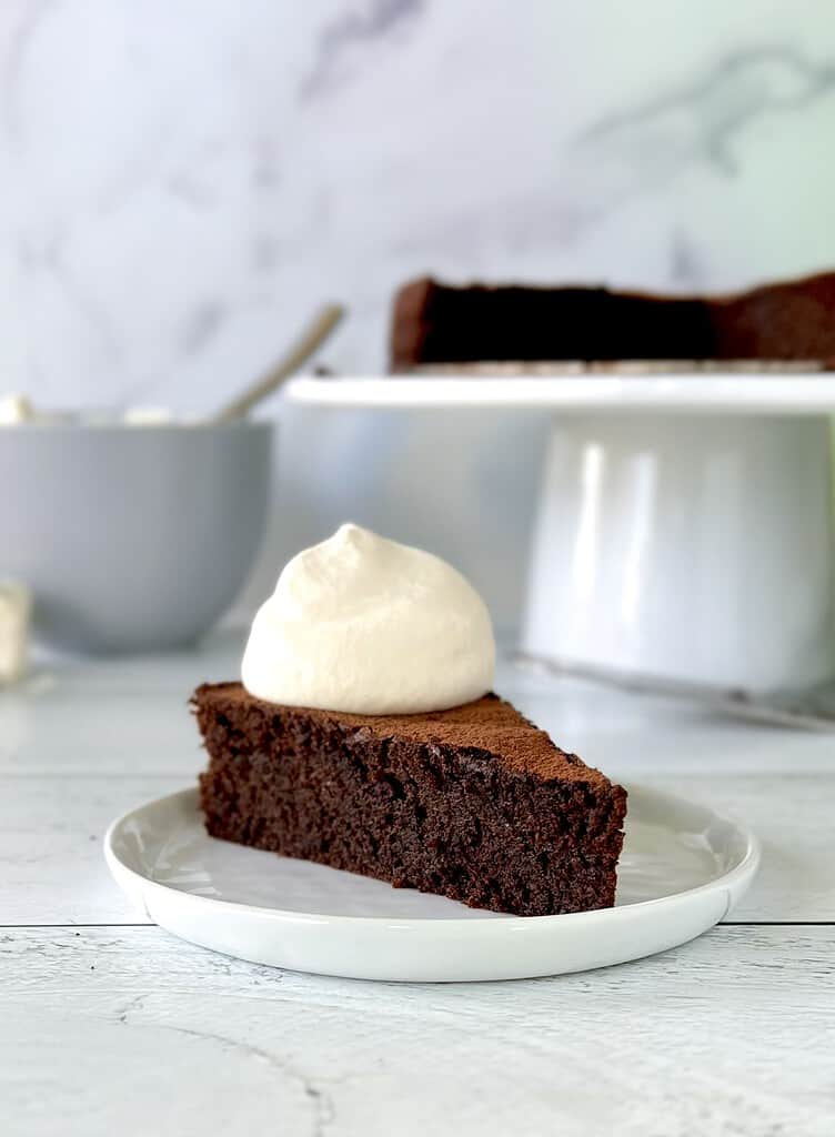 A slice of flourless dairy free chocolate cake topped with whipped cream on a white plate.
