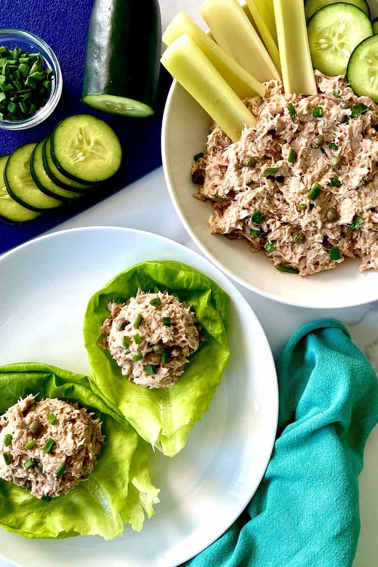 Healthy tuna salad with Caesar dressing in a bowl with celery sticks and sliced cucumbers and on a plate scooped into lettuce cups.