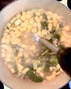A stick blender blending apples, broccoli, onions and almonds in water in a soup pot.