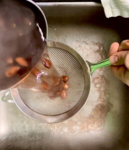 Draining boiled almonds through a sieve over a sink.
