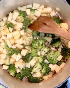 A wooden spatula stirring broccoli, almonds, onions and apple in water a soup pot.