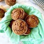Paleo banana muffins in a towel-lined bowl next to more muffins on a marble table.