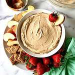 Peanut butter and yogurt dip in a bowl on a plate surrounded by apple slices, graham crackers and strawberries.