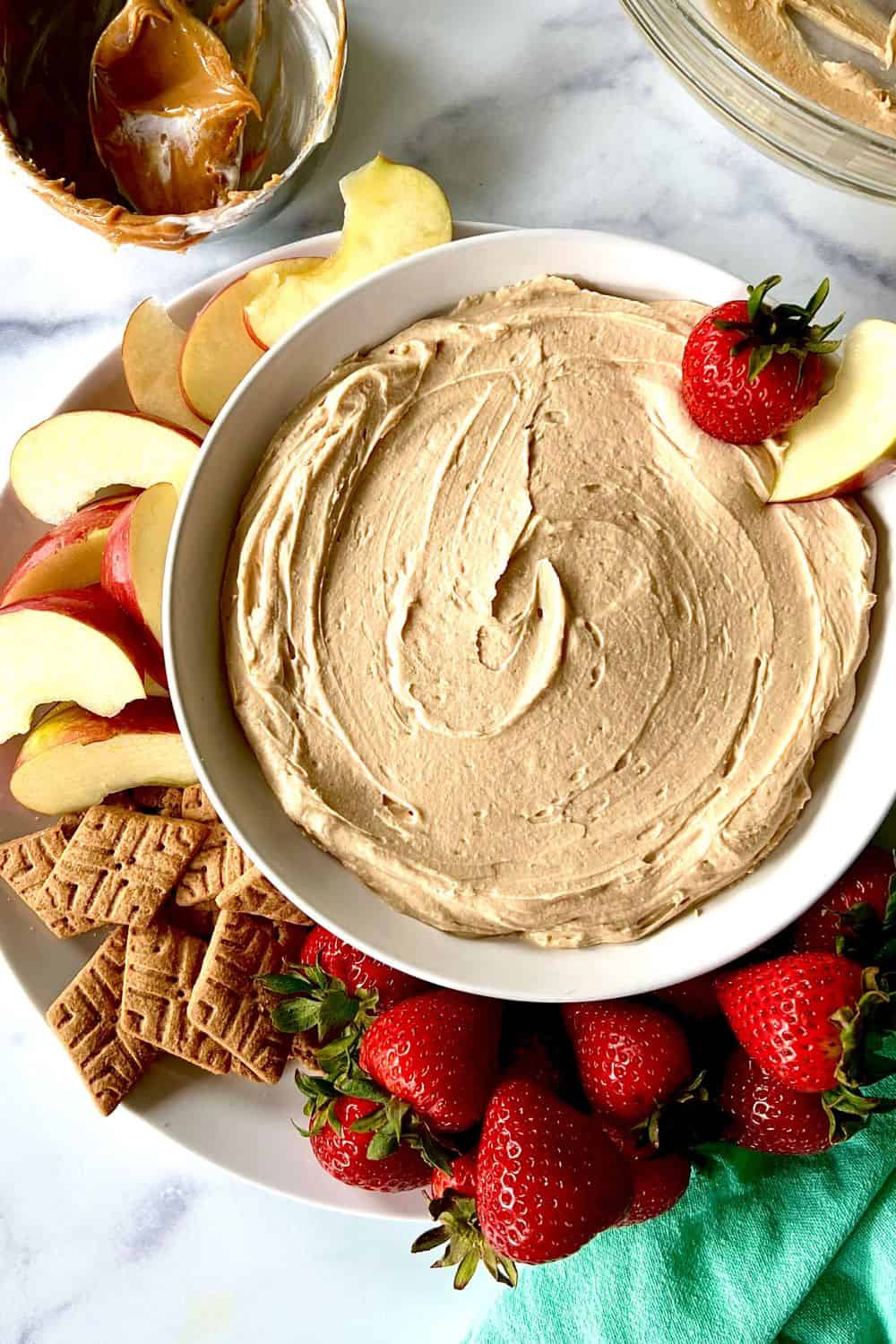 Peanut butter yogurt dip in a bowl on a plate surrounded by strawberries, apple slices and graham crackers.