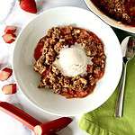 Healthy strawberry rhubarb crisp in a bowl topped with vanilla ice cream, next to a napkin, spoon, strawberries and stalks of raw rhubarb.