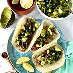 A Paleo ground bison recipe for tacos in tortillas topped with chunky avocado and blueberries on a plate with lime wedges.