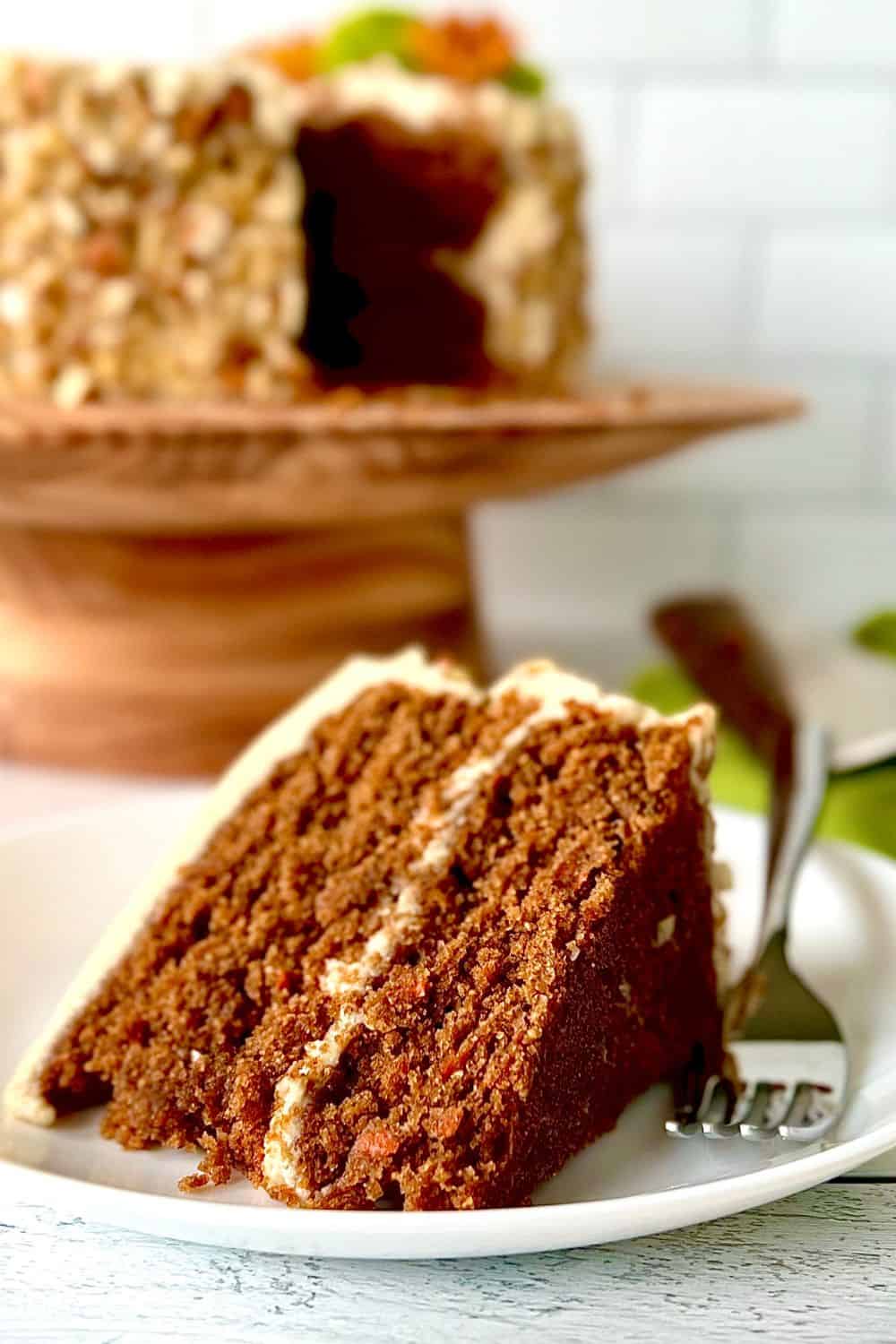 A slice of gluten-free carrot cake on a plate with the whole cake in the background on a cake stand.