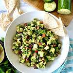 Avocado and cucumber salsa in a bowl next to tortilla chips, sliced cucumber, fresh parsley and a towel.