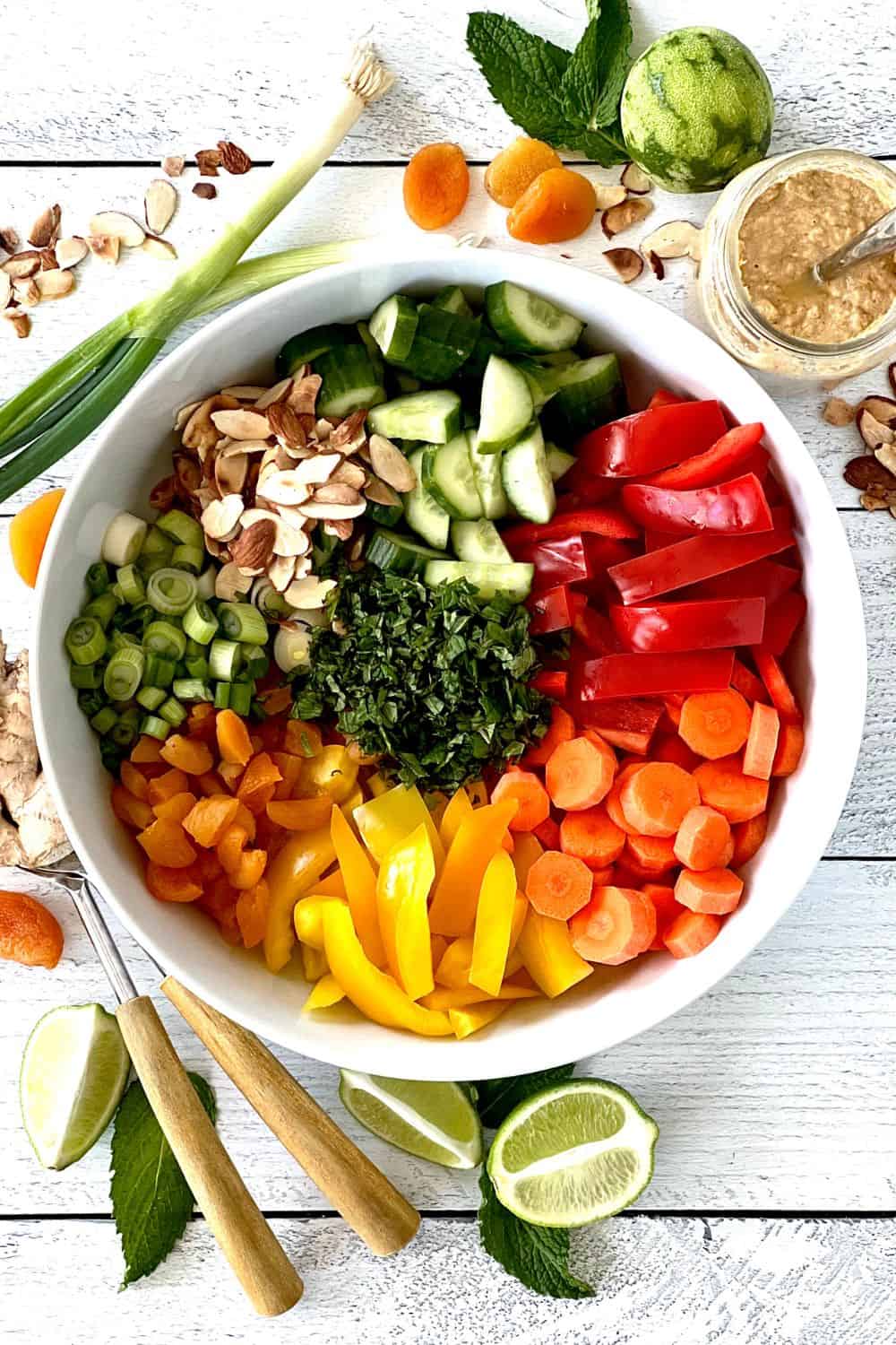 A colorful salad with peppers, herbs, sliced almonds, cucumber, carrots, dried apricots and green onions in a large bowl.