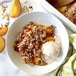 Peach crisp without oats in a bowl with a scoop of vanilla ice cream, next to peach slices, sliced almonds and a striped towel with a spoon on it.