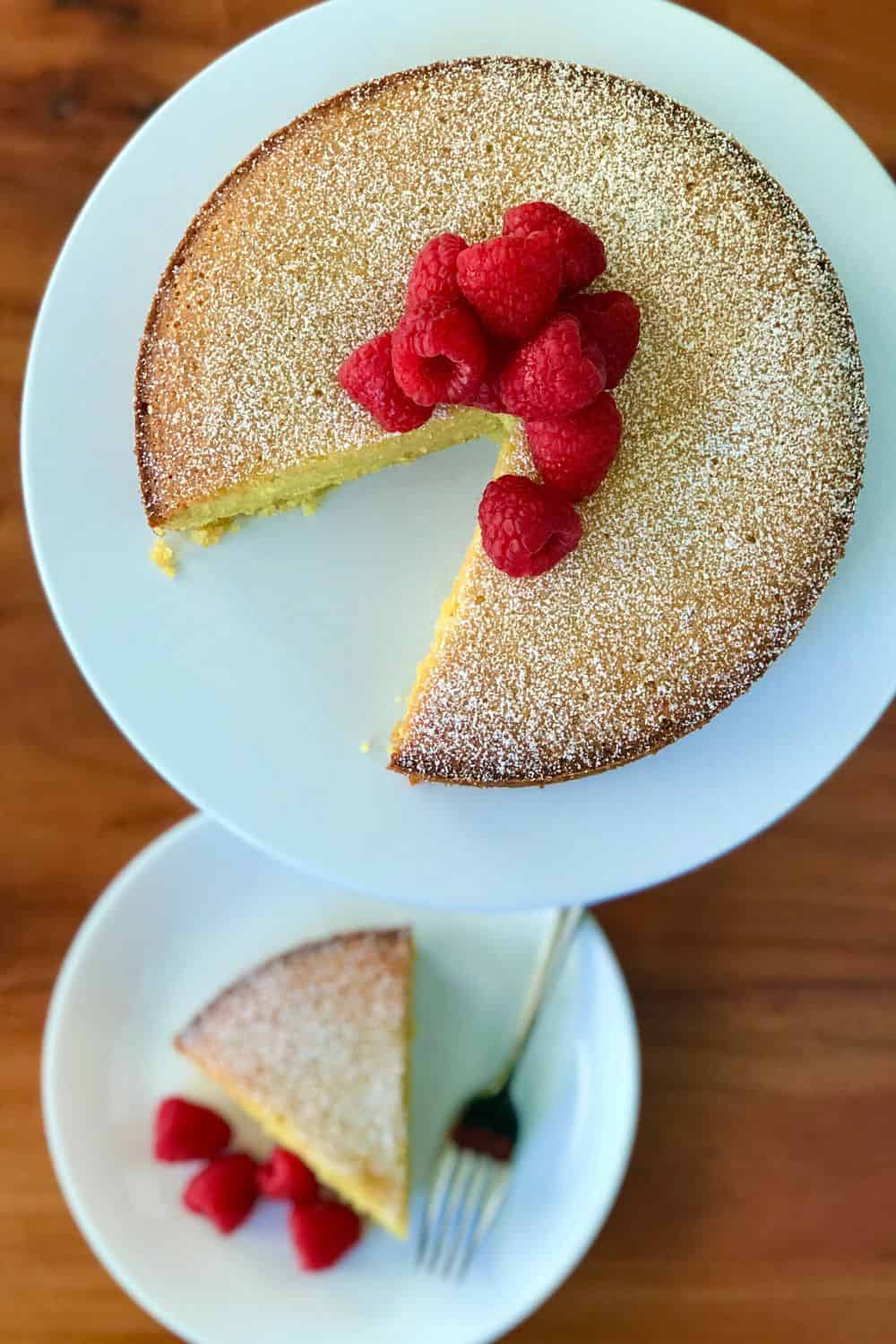 Olive Oil Polenta Cake topped with raspberries on a cake stand and a slice of cake and raspberries on a plate next to it.