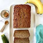 A loaf of healthy zucchini banana bread and 2 slices on a platter next to bananas and zucchini.