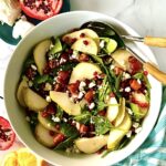 A pear and pomegranate salad in a large serving bowl with 2 salad servers.