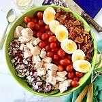 Healthy cobb salad in a large bowl with rows of diced red onion, chicken, grape tomatoes, hard boiled eggs and chopped bacon.