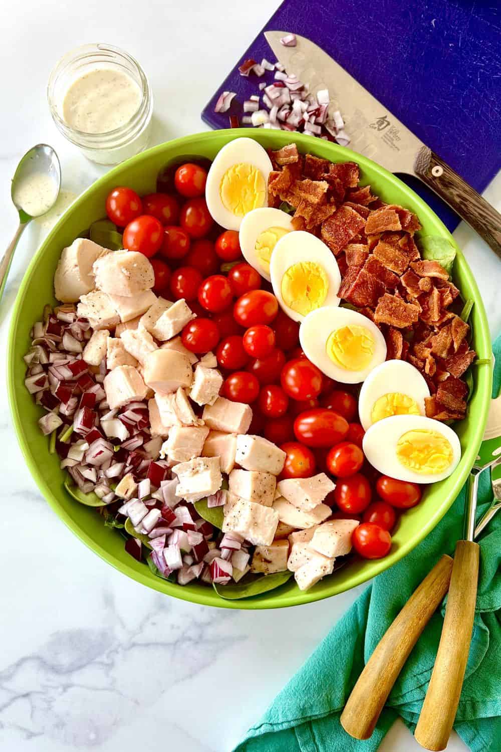 Whole30 cobb salad in a large bowl with rows of diced red onion, chicken, grape tomatoes, hard boiled eggs and chopped bacon.