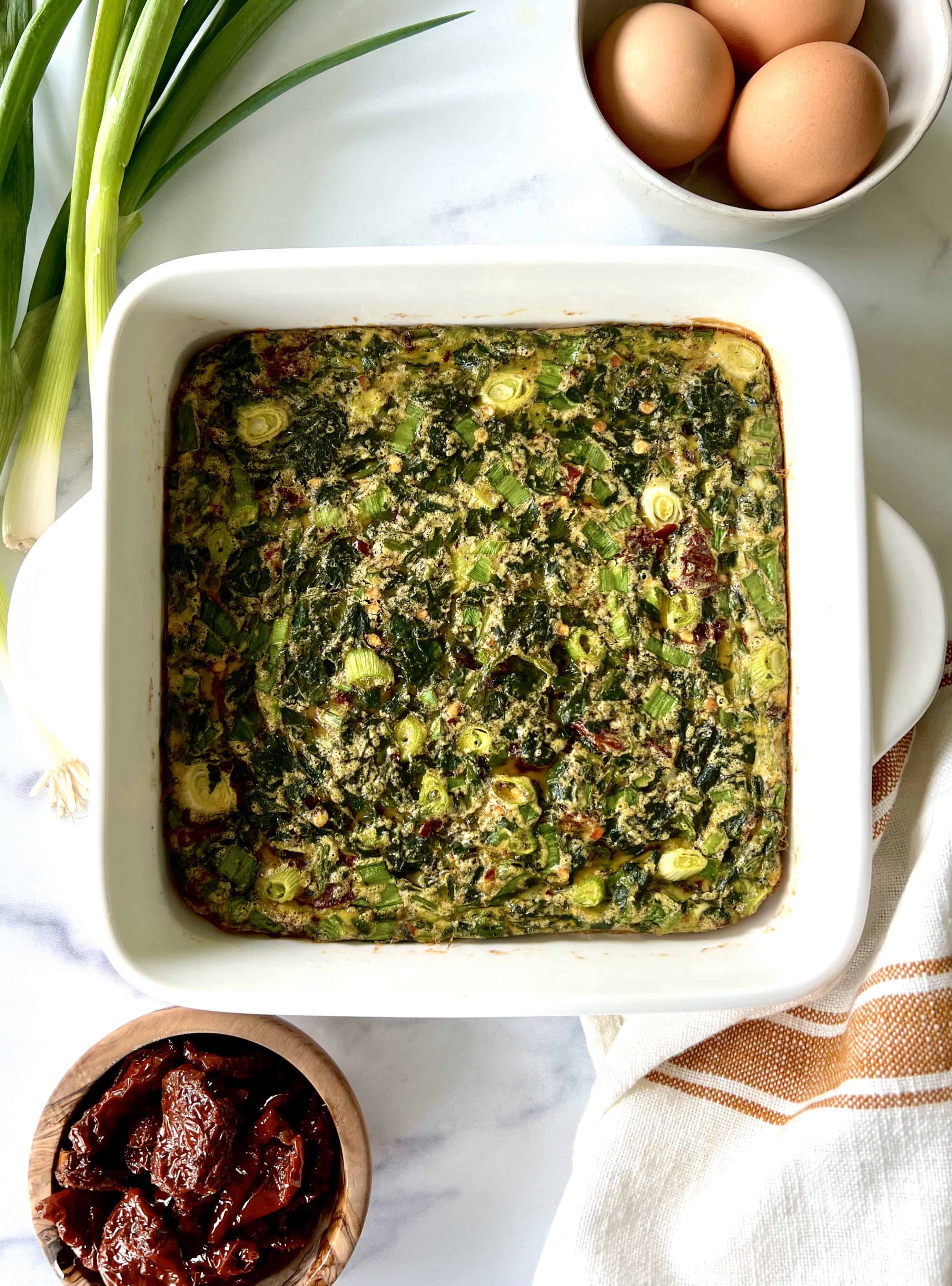A keto spinach casserole in a square baking dish next to bowls of eggs and sun-dried tomatoes.