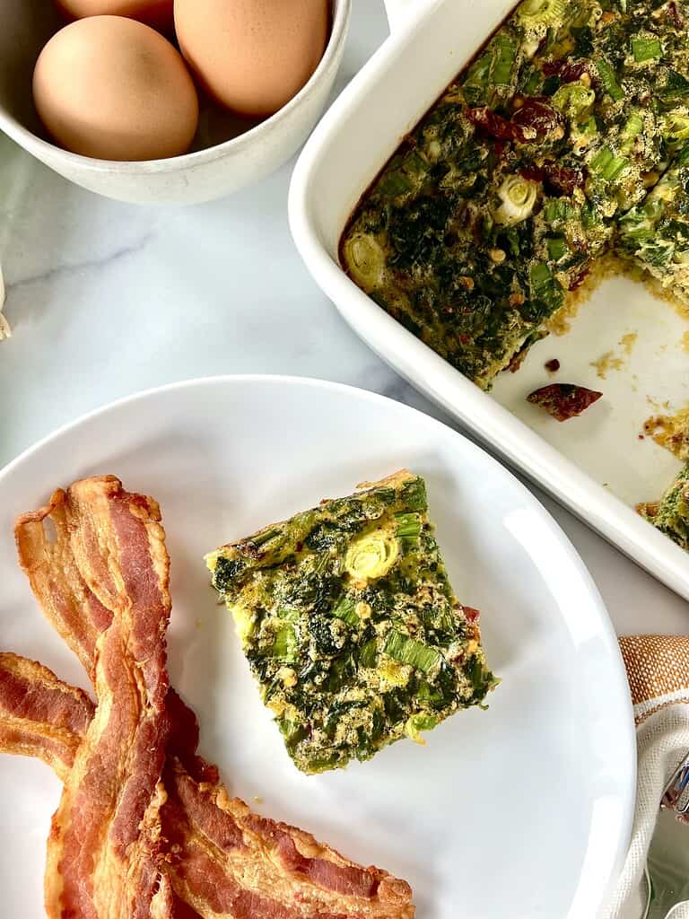 A square slice of an egg and spinach bake on a plate with bacon next to more of the egg bake in a square baking dish.