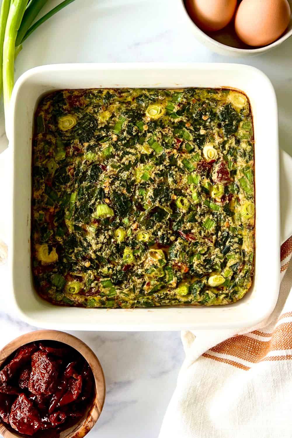 Dairy free egg bake in a square baking dish next to a towel and a bowl of sun-dried tomatoes.