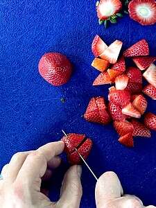 Two hands using a paring knife to chop strawberries on a cutting board.