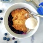 Dairy-free blueberry cobbler in a bowl with a spoon and a scoop of vanilla dairy-free ice cream.