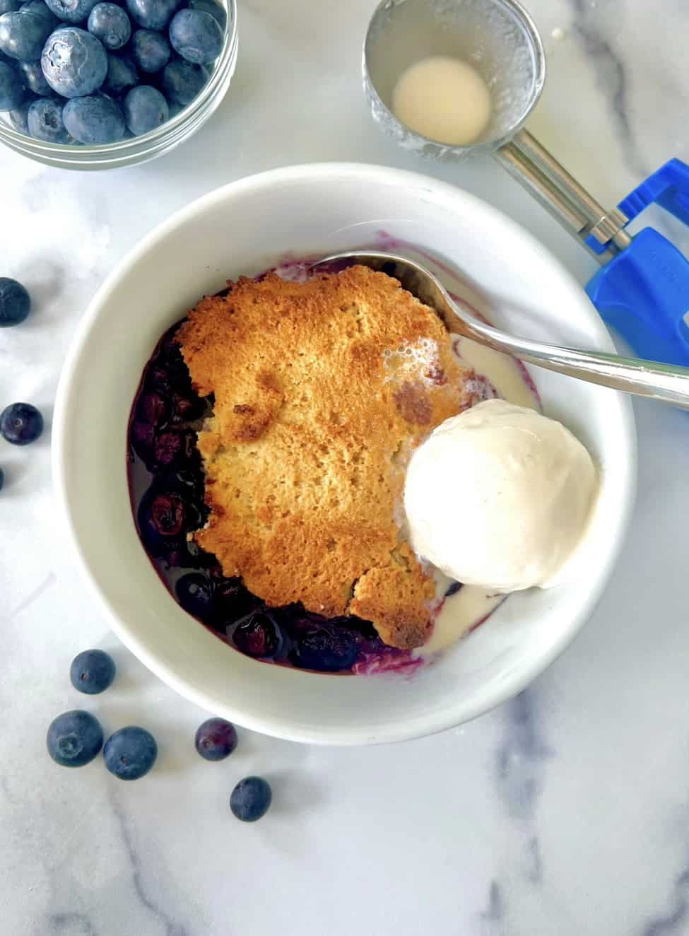 Dairy-free blueberry cobbler in a bowl with a spoon and a scoop of vanilla dairy-free ice cream.