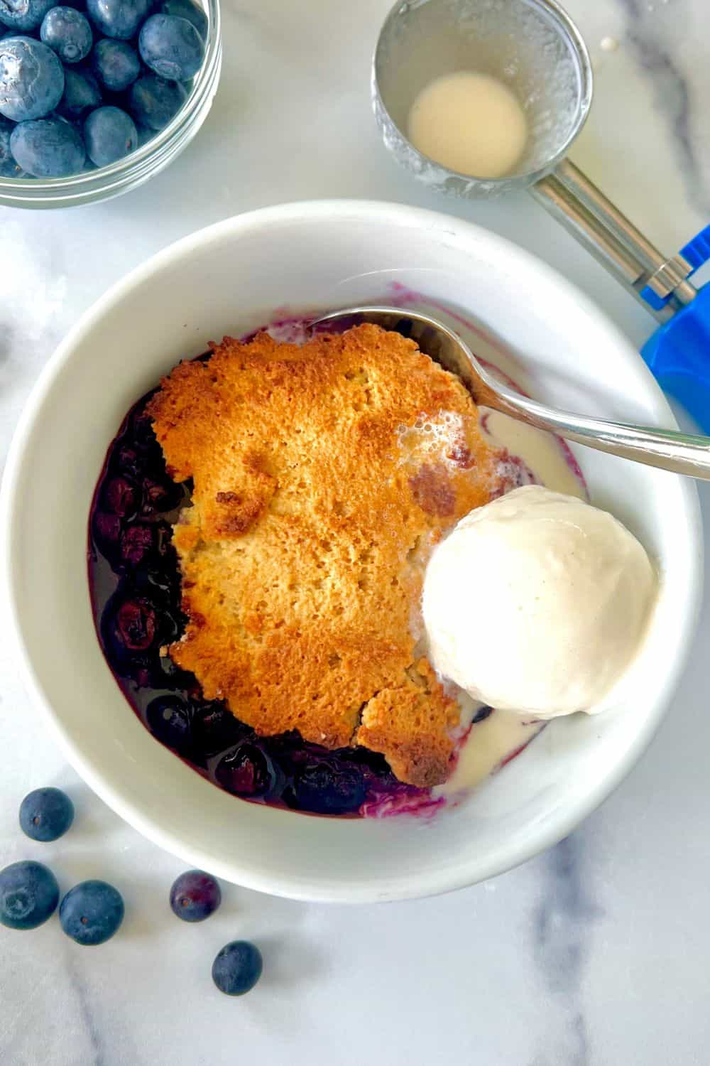Gluten free blueberry cobbler in a bowl with a spoon and scoop of vanilla ice cream.
