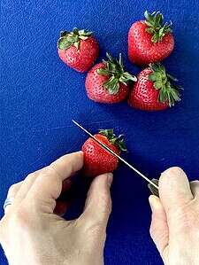 Two hands using a paring knife to hull strawberries on a cutting board.