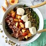 Burrata and arugula salad arranged in a bowl with chopped peaches, torn prosciutto and pistachios.