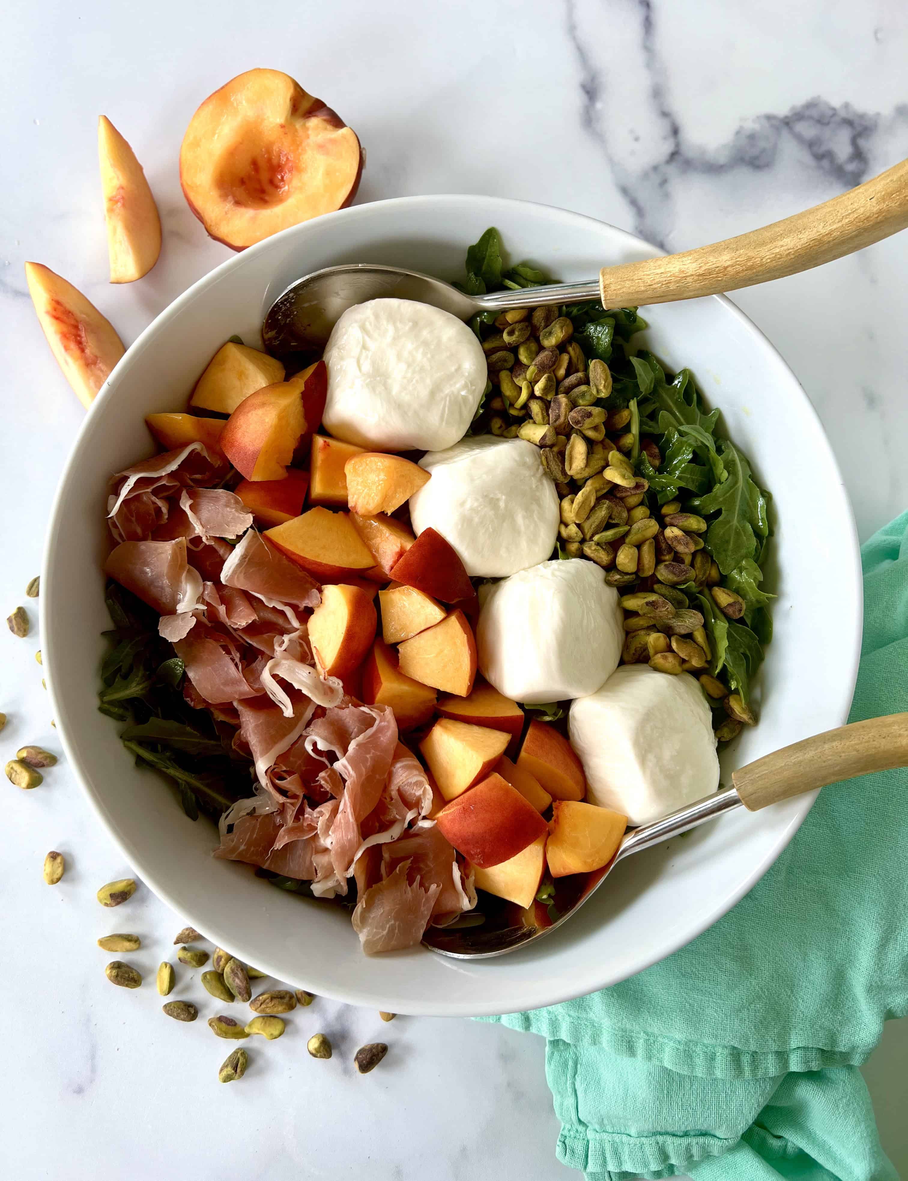 Burrata and arugula salad arranged in a bowl with chopped peaches, torn prosciutto and pistachios.