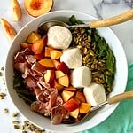 An arugula and burrata salad arranged in a large bowl with pistachios, chopped peaches and torn prosciutto.