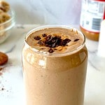 A Chocolate Peanut Butter Banana Smoothie in a tall glass topped with a peanut butter drizzle and bits of chocolate.