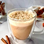 A gluten free pumpkin spice latte in a glass mug next to two cinnamon sticks.