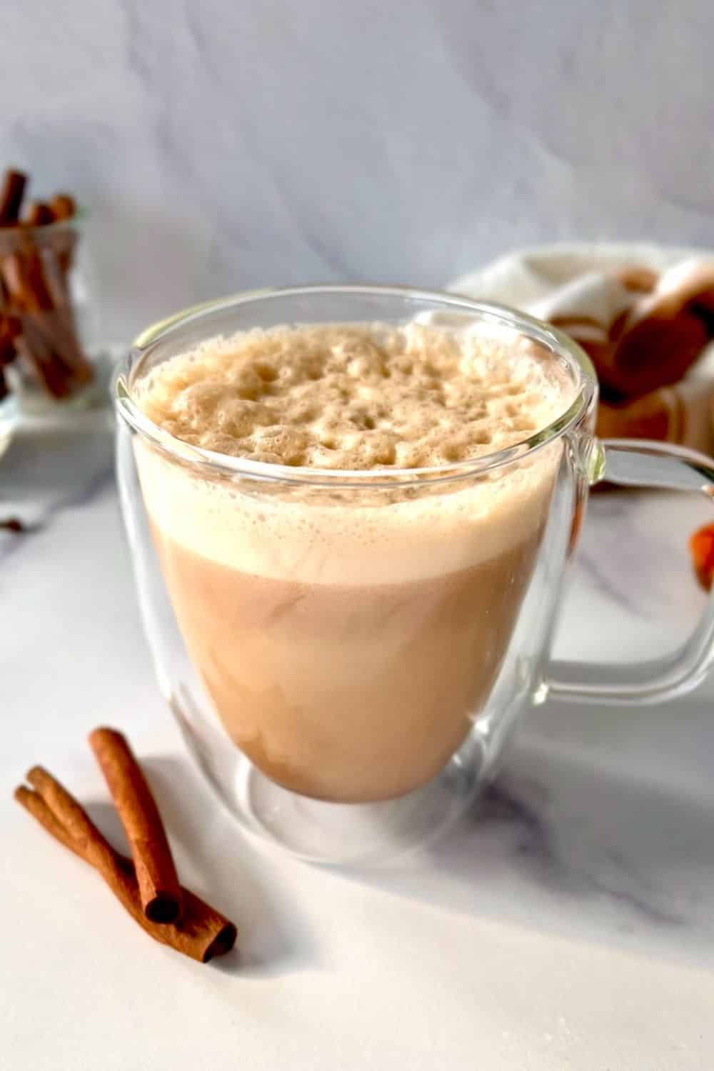 A gluten free pumpkin spice latte in a glass mug next to two cinnamon sticks.